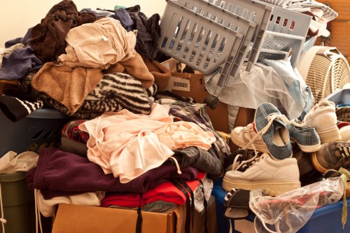 A large pile of mixed household waste, including clothing, textiles, and fabric items such as sweaters, t-shirts, and towels, all in various colours like beige, brown, red, and pink, is shown piled on a flat surface. The materials appear crumpled and layered, with some garments draping over the edges of cardboard boxes and plastic containers. Among the clothing heap are a pair of light-colored sneakers and a plastic bag containing more items, partially visible at the bottom right. The setting appears to be an indoor or garage environment with a wooden surface and a plastic laundry basket, indicating collection for rubbish removal. The overall scene demonstrates the kind of clutter stockpiled for disposal or clearance, typical of rubbish collection tasks performed by a waste management service such as Commercial Waste Guildford, serving areas in and around the Guildford postcode region.