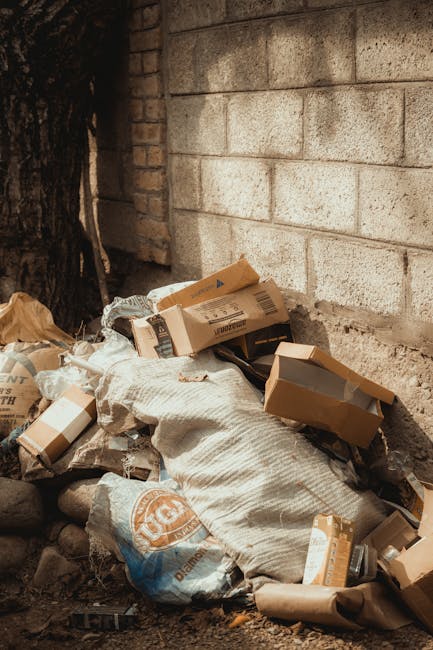 A pile of discarded household waste and debris is situated outdoors on a paved surface, near a brick wall. The collection includes wooden slatted furniture that appears weathered and partly broken, wi