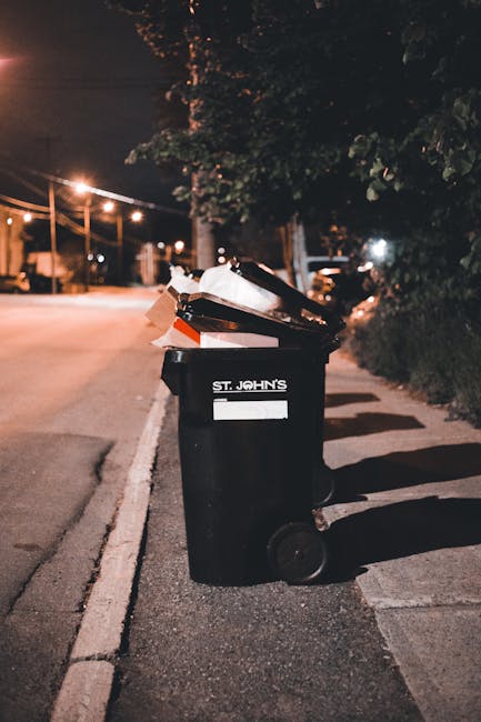A large collection of overflowing rubbish bins and scattered waste bags, paper and cardboard boxes, and household waste spill onto the pavement in front of a commercial building with storefronts. The 