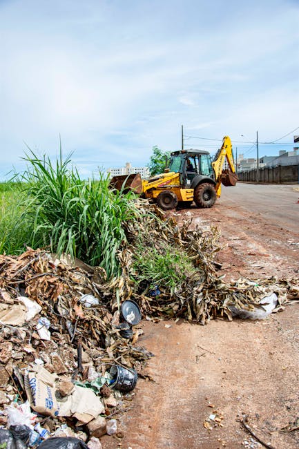 A collection of various discarded rubbish and waste materials piled on a concrete driveway, including large cardboard boxes, plastic bags filled with debris, and assorted broken or unused items with r